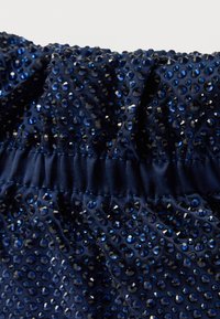 Tmavomodrá tkanina s textúrovaným povrchom, ozdobená mnohými lesklými modrými kamienkami, s nariaseným, elastickým pásom.