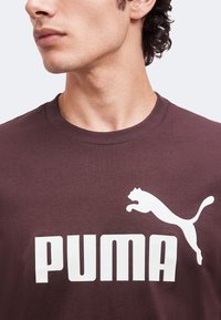 Νέος άνδρας που φοράει ένα σκούρο καφέ t-shirt Puma με λευκό λογότυπο Puma και κείμενο, κοιτάζει στο πλάι μπροστά σε ένα απλό φόντο.