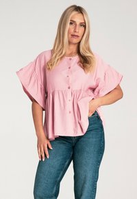 Różowa koszula zapinana na guziki z krótkimi, szerokimi rękawami i zgromadzonym w pasie materiałem. Noszona z niebieskimi dżinsami, prezentowana na neutralnym tle.
