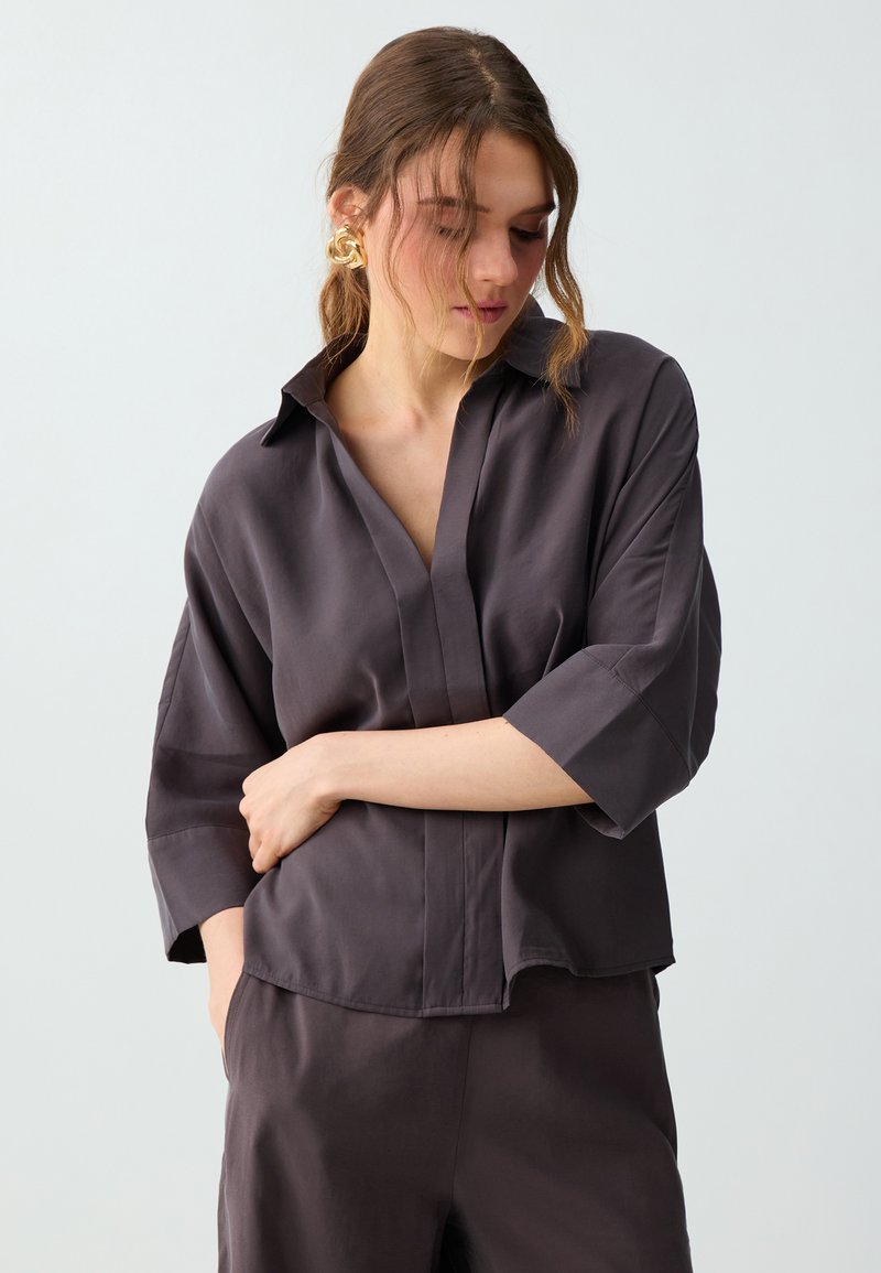 Femme aux cheveux bruns portant des boucles d'oreilles en or, une chemise ample gris foncé à boutons avec manches trois-quarts et un pantalon assorti, regardant vers le bas.