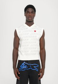 Hvid ærmeløs sweater med gentaget "ICECREAM" mønster, parret med sorte bukser med en blå hundegrafik på siden. Rød logoaccent.