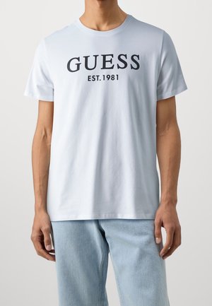 Mann trägt weißes T-Shirt mit schwarzem Aufdruck "GUESS EST. 1981", kombiniert mit hellblauen Jeans, gezeigt von Hals bis Hüfte.