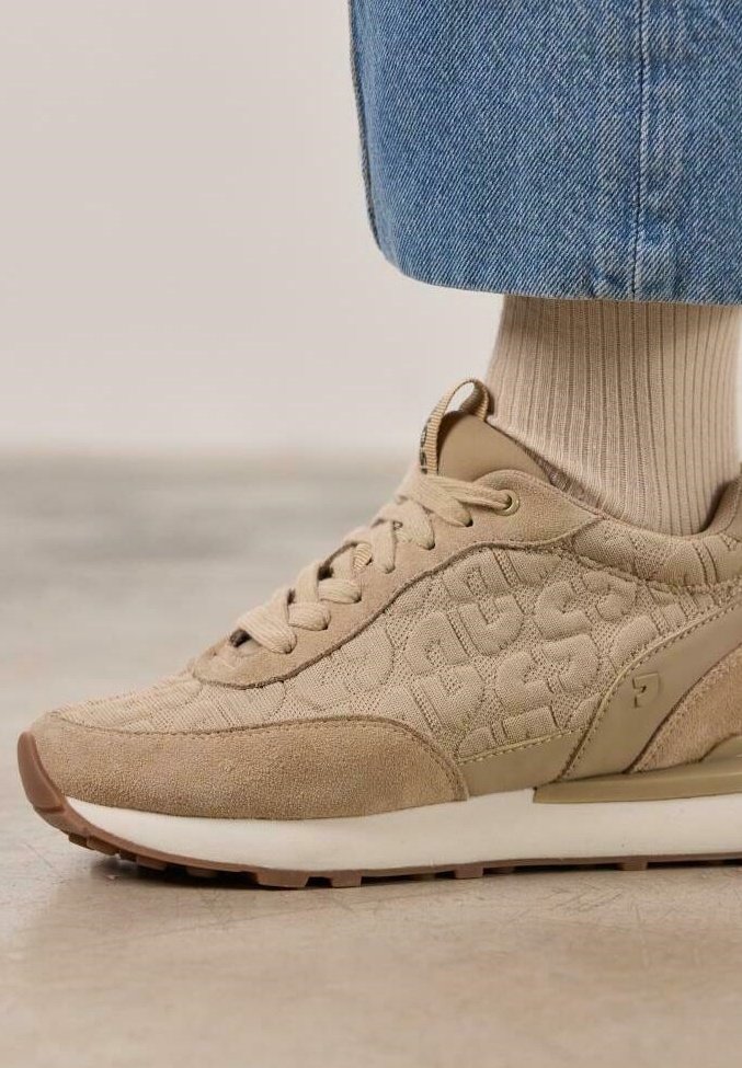 Zapatilla beige con parte superior texturizada, que presenta un diseño estampado, cierre con cordones y una suela blanca. Detalles en un tono ligeramente más oscuro.