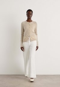 Beige cardigan med knappar, bärs över vita utsvängda byxor. Modellen står mot en enfärgad bakgrund och visar upp den lätta stickade texturen.