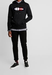 Svart hoodie med en central logga i rött, vitt och marinblått; lager-på-lager över en svartvit rutig skjorta; kombinerat med svarta byxor och svarta sneakers.