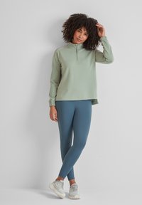 Light green zip-up pullover with long sleeves and ribbed cuffs, paired with high-waisted teal leggings and white sneakers. Smooth texture.