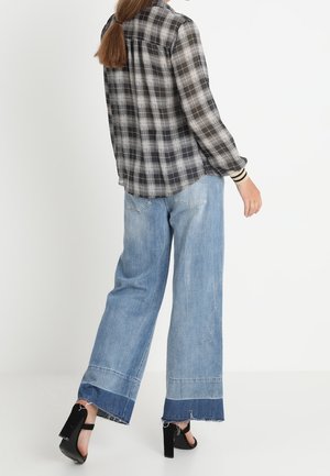 Personne portant un jean bleu clair à jambes larges avec des ourlets foncés, des sandales noires à talons hauts et une chemise à carreaux noire et blanche.