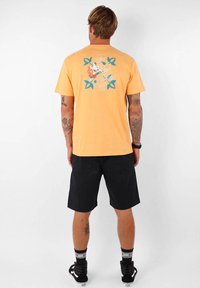 T-shirt pour homme à manches courtes, orange, avec un imprimé graphique floral en vert et blanc au dos. Associé à un short noir et des chaussures noires.
