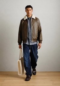 Giubbotto bomber in pelle marrone con colletto in shearling, camicia a quadri blu, jeans scuri e scarpe marroni, che porta una borsa tote beige.