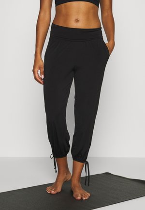 Femme debout pieds nus sur un tapis de yoga noir, portant un pantalon noir à cordon aux chevilles et un haut court noir.