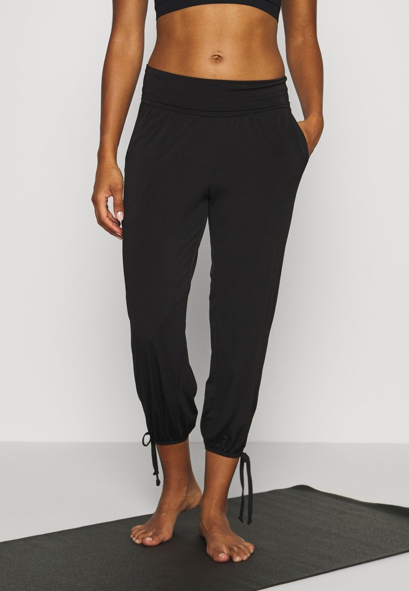 Femme debout pieds nus sur un tapis de yoga noir, portant un pantalon noir à cordon aux chevilles et un haut court noir.
