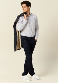 Witte overhemd met kleine blauwe stippen, donkere denim jeans en witte sneakers. Zwarte jas over de schouder gedrapeerd, gladde textuur.