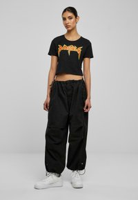 Tricou scurt negru cu grafică "BAD GIRLS" în roșu și galben, asociat cu pantaloni cargo negri, cu croială largă, și teniși albi.