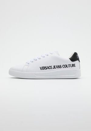 Weißer Sneaker mit schwarzem Fersendetail, weißen Schnürsenkeln und "Versace Jeans Couture", das in fetten schwarzen Buchstaben seitlich aufgedruckt ist.