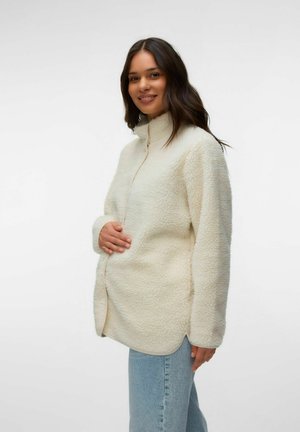 Femme souriante aux longs cheveux foncés portant une veste polaire crème et un jean bleu clair, tenant doucement son côté.