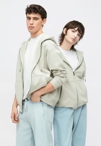 Du modeli dėvi šviesiai pilkus užtrauktuku užsegamus džemperius su kapišonu, kuriais apsivilkę baltus marškinėlius ir derina su šviesiai mėlynais džinsais. Džemperiai turi šonines kišenes ir ribotuotas rankogalius.