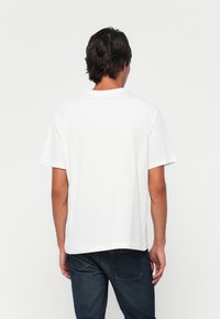 Mand med mørkt hår iført en ensfarvet hvid kortærmet t-shirt og mørkeblå jeans, som står med ryggen til mod en ensfarvet baggrund.