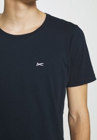 Námořnická modrá bavlněná tričko s kulatým výstřihem; má malé bílé logo nůžek na levém prsu a krátké rukávy.