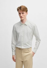 Helle, knopfbare Bluse mit einer subtilen strukturierten Textur und kleinen blauen Akzenten, kombiniert mit beigen Hosen.