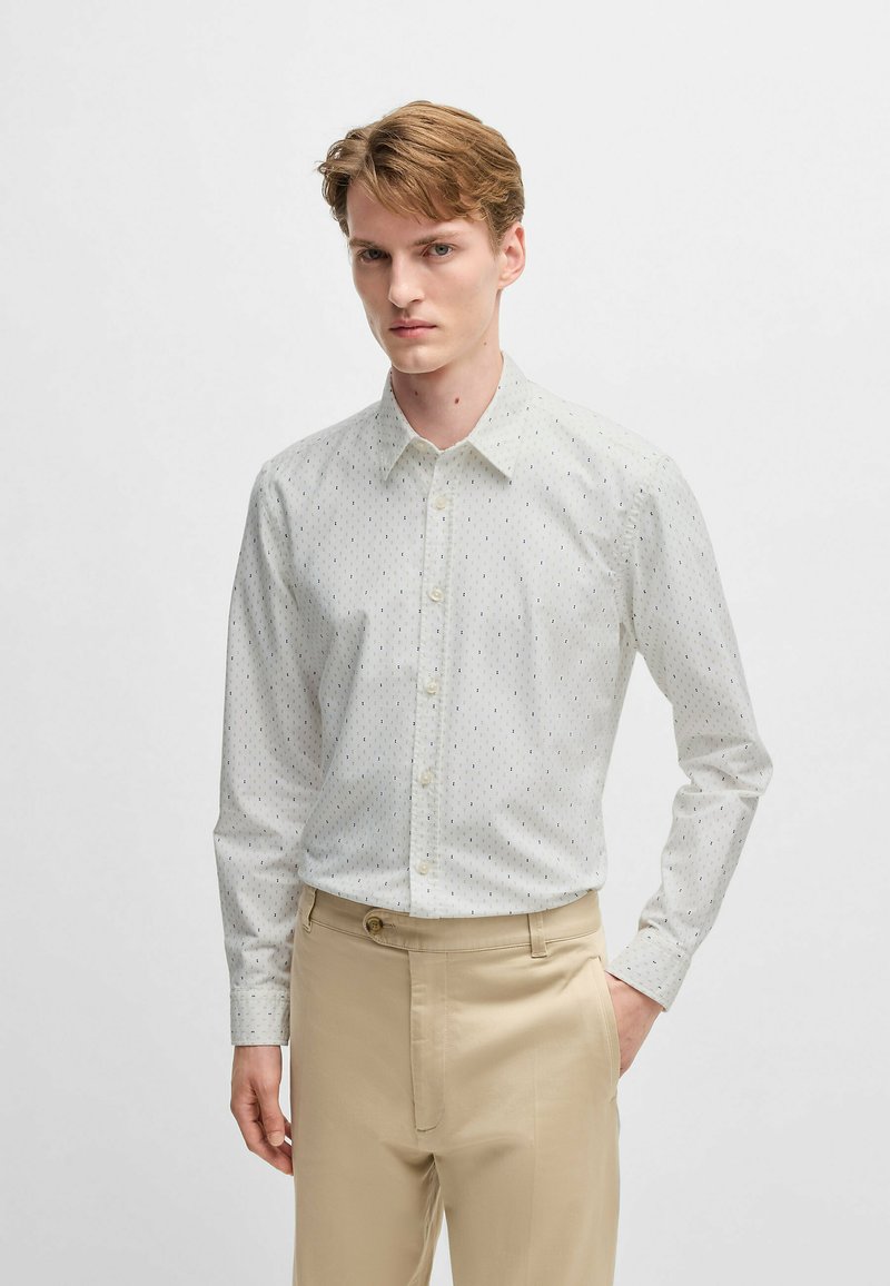 Lichtgekleurde overhemd met een subtiele gestructureerde textuur en kleine blauwe accenten, gecombineerd met beige broeken.
