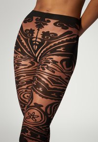 Sorte leggings med mønster og gennemsigtigt stof, der har detaljerede svulster og blomsterdesigns, med et solidt sort bånd i taljen for støtte.