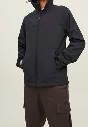 Personne portant une veste noire zippée avec les mains dans les poches avant et un pantalon cargo marron sur un fond uni.