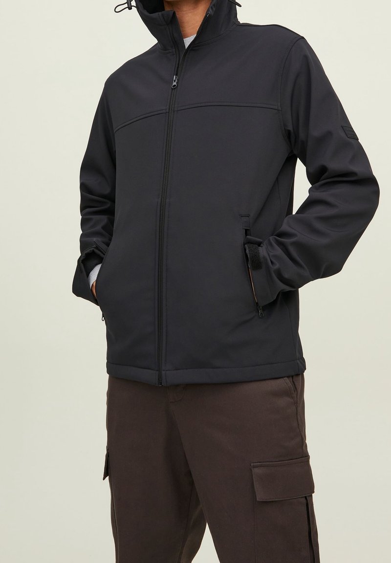 Personne portant une veste noire zippée avec les mains dans les poches avant et un pantalon cargo marron sur un fond uni.
