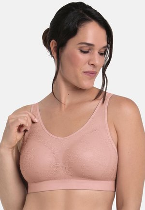 Woman wearing a light pink lace patterned soft bra, holding one strap, with dark hair tied back against a plain light background.