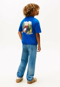 T-shirt in cotone blu con una grafica colorata di campeggio sul retro, abbinata a jeans in denim azzurro chiaro e sneakers beige.