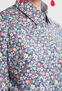 Chemise à boutons avec un fond bleu foncé et un motif floral en rose, blanc et vert. Col avec une texture lisse.