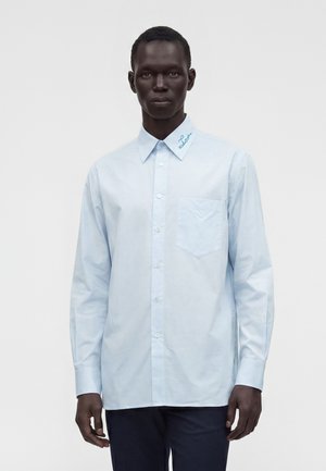 Homme portant une chemise bleu clair à manches longues avec une poche poitrine et un texte brodé sur le col, debout devant un fond uni.