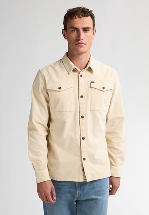 Light beige button-up shirt made of textured fabric, featuring two chest pockets, brown buttons, and a small logo tag on the left pocket.