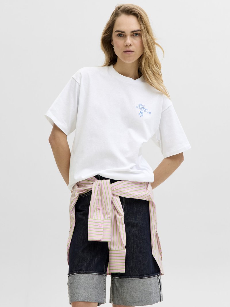 Νεαρή γυναίκα με μακριά ξανθά μαλλιά, που φοράει λευκό μπλουζάκι, σκούρα σκισμένα τζιν σορτς και ριγέ πουκάμισο δεμένο γύρω από τη μέση, στέκεται μπροστά σε λευκό φόντο.