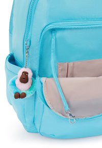 Turkos ryggsäck med en slät, glänsande yta, som har en mjuk apa nyckelring och en framlapp med en beige insida.