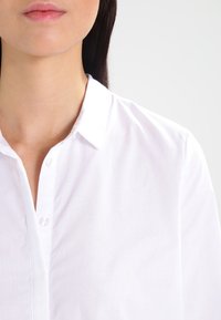 Vit blus med krage, med en knappstängning fram. Tyget ser slätt och något genomskinligt ut.