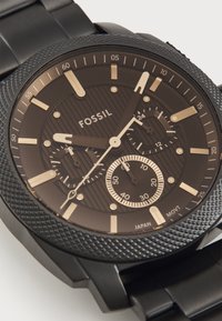 Montre Fossil avec un boîtier en acier inoxydable noir, une lunette texturée, un cadran sombre avec des accents dorés, des sous-cadrans et des index.