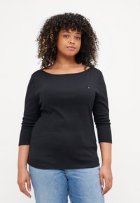 Svart langermet topp med båthals, med en liten logo på venstre bryst. Myk, elastisk stoff kombinert med lyseblå jeans.
