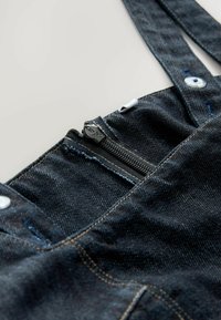 Salopette en denim avec tissu noir, dotée d'une poche avant zippée, de fermetures à boutons et d'accents de couture bleus. Bretelles ajustables.