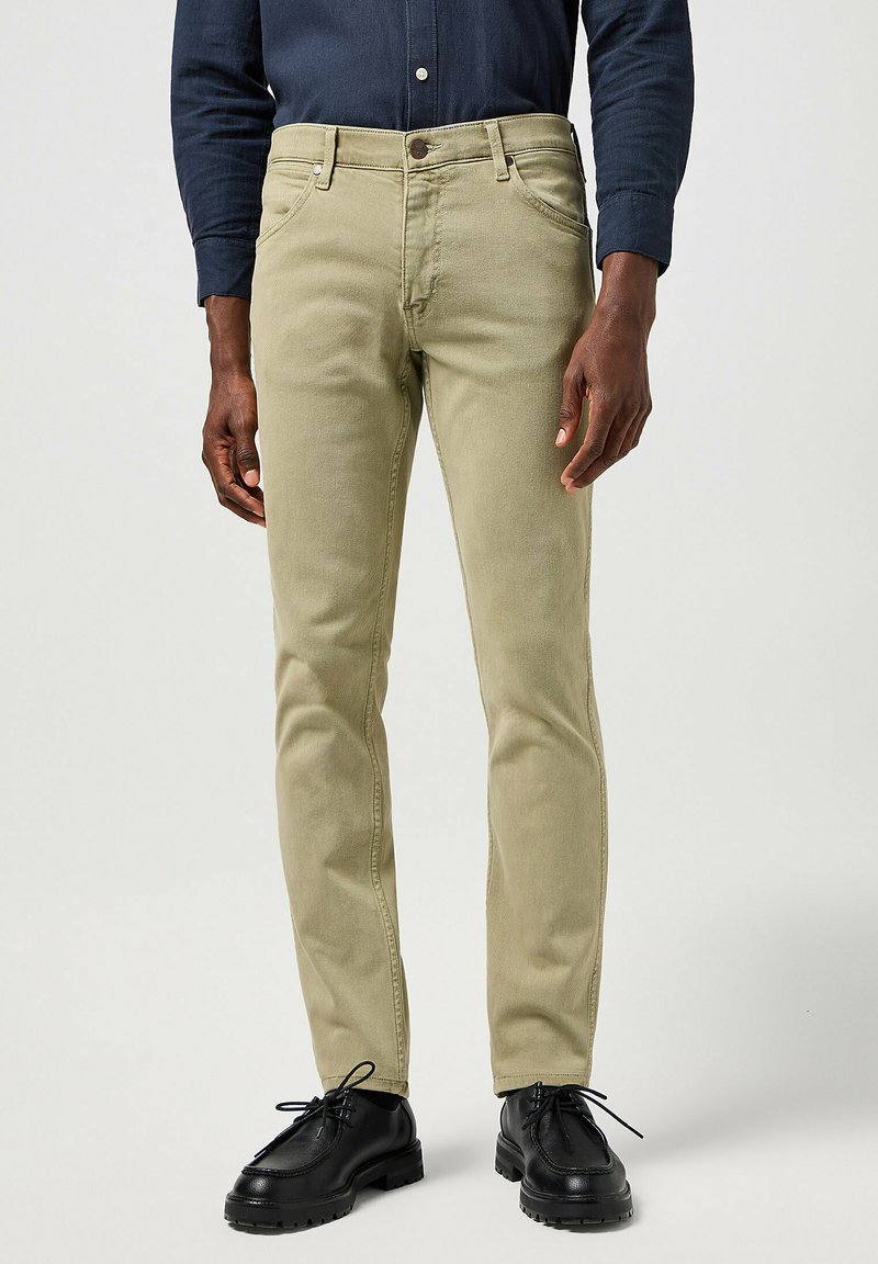 Pantalones de algodón color caqui con un corte slim y frente plano, con cinco bolsillos y cierre de un botón. Llevados con zapatos negros de cordones.