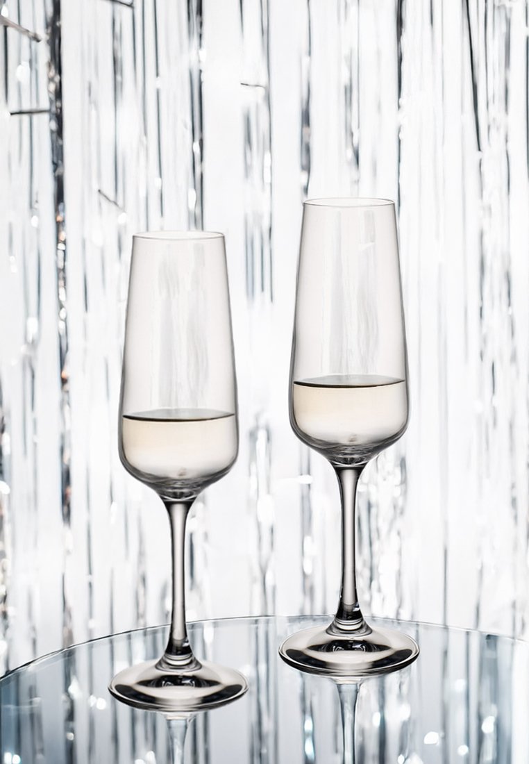 Dwa przezroczyste kieliszki do szampana z cienkimi nóżkami, częściowo napełnione jasnym płynem, ustawione na lustrzanej powierzchni na tle błyszczącego srebrnego tła.