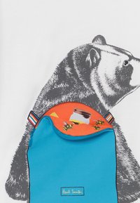 Modrá taštička se zipem, uvnitř s oranžovým vzorem a včelami. Překrývá bílou košili s šedým grafickým designem medvěda.