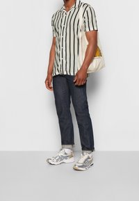 Gestreept shirt met korte mouwen in crème en zwart, gecombineerd met donkere spijkerbroek en zilveren sportschoenen; draagt een beige draagtas met gouden accenten.