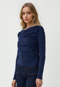 Maglia a maniche lunghe in marino con scollo drappeggiato, arricciature laterali e texture del tessuto liscia; abbinata a jeans scuri con tasche posteriori.