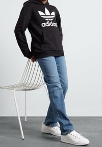Sudadera negra con capucha y logo blanco de Adidas, jeans azules y zapatillas blancas, de pie junto a una silla de metal blanca contra un fondo neutro.