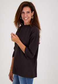 Donkerbruin pullover met een hoge halslijn, driekwart mouwen en een relaxte pasvorm, gecombineerd met lichtblauwe denim jeans.