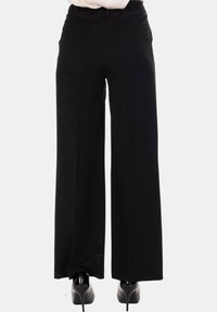 Donna che indossa pantaloni neri a gamba larga e scarpe nere con tacco alto, in piedi davanti a uno sfondo bianco uniforme.