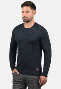 Dark blue, long-sleeved sweater made of knit fabric. Features a round neckline and ribbed hem, with a small brand patch at the waist.