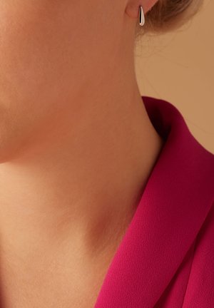 Des boucles d'oreilles en argent pendent de l'oreille, présentant une forme de goutte lisse et allongée. La porteuse porte un haut rose à la texture douce.