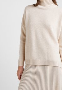 Pull en tricot beige avec un col roulé côtelé haut et des épaules tombantes, doté d'un ourlet côtelé, porté avec une jupe plissée.