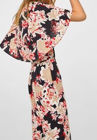 Květinový overal s velkými, volnými rukávy, přiléhavým pasem a kombinací červené, béžové a černé barvy v odvážném vzoru na pevném pozadí.